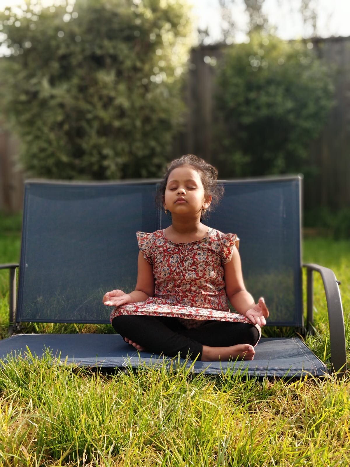 Child sitting cross-legged on bench, meditating outdoors in sunny garden