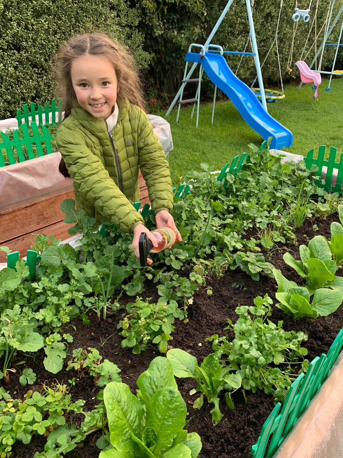 Young girl in a green jacket holding a spray bottle, tending to plants in a garden with a playground slide in the background.