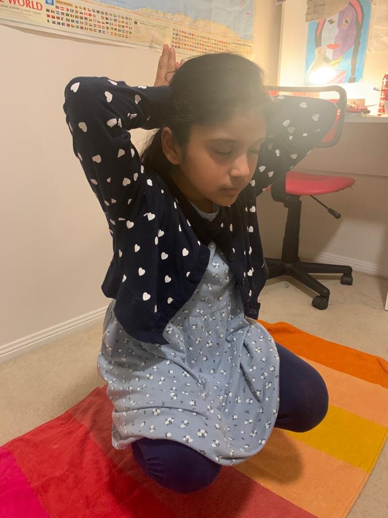 Child practicing yoga indoors, kneeling on a colorful mat, with eyes closed and hands on her head. Room has a desk and chair.