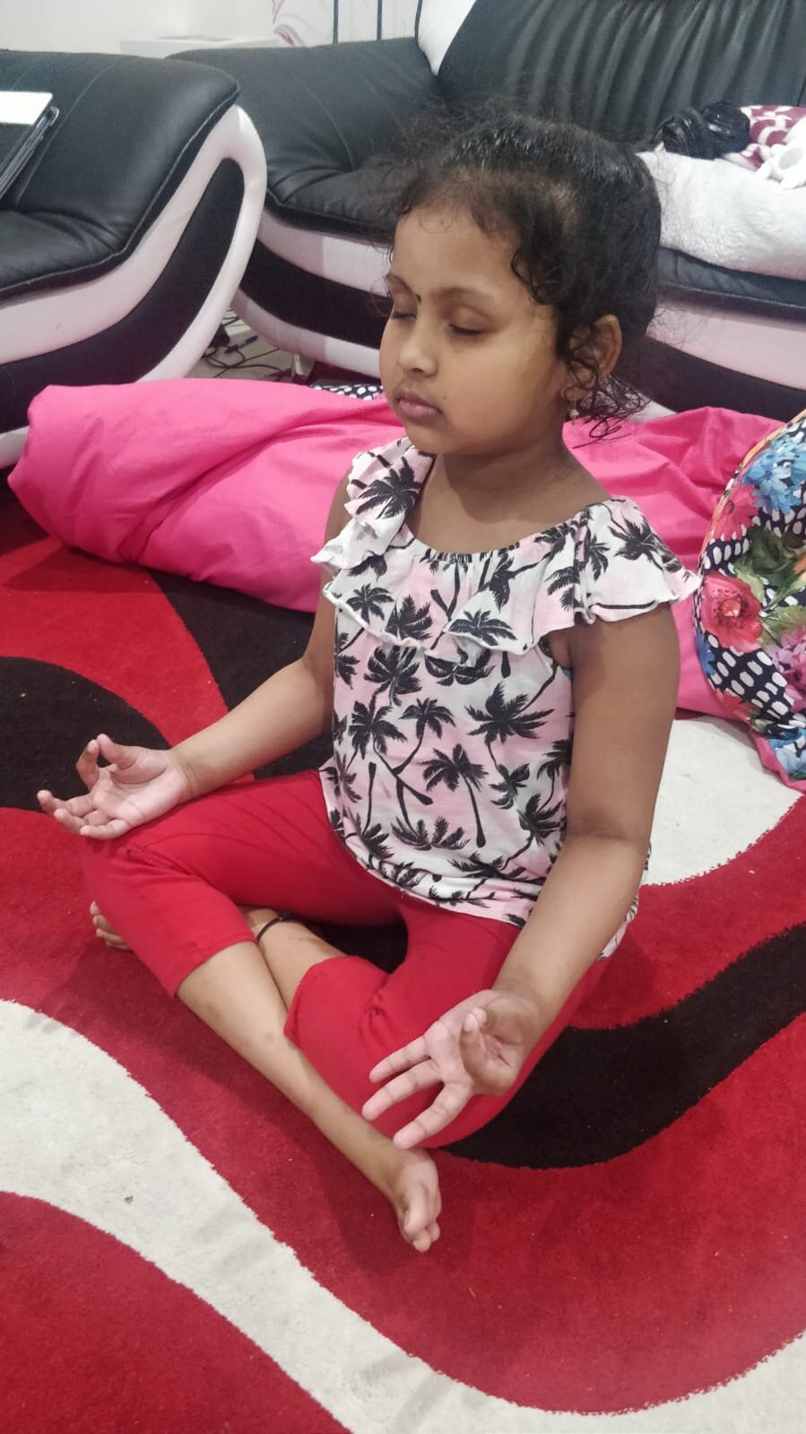 A young child sitting cross-legged on a red and black patterned rug, meditating with eyes closed. The child is wearing a palm tree print top and red pants. In the background, there is a black and white leather sofa with cushions.