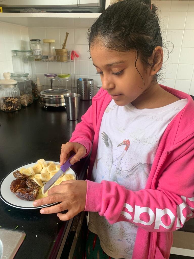 Girl in pink jacket cutting a banana on a plate in a kitchen.