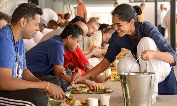 Group of people sitting and being served food on leaf plates in a communal setting.