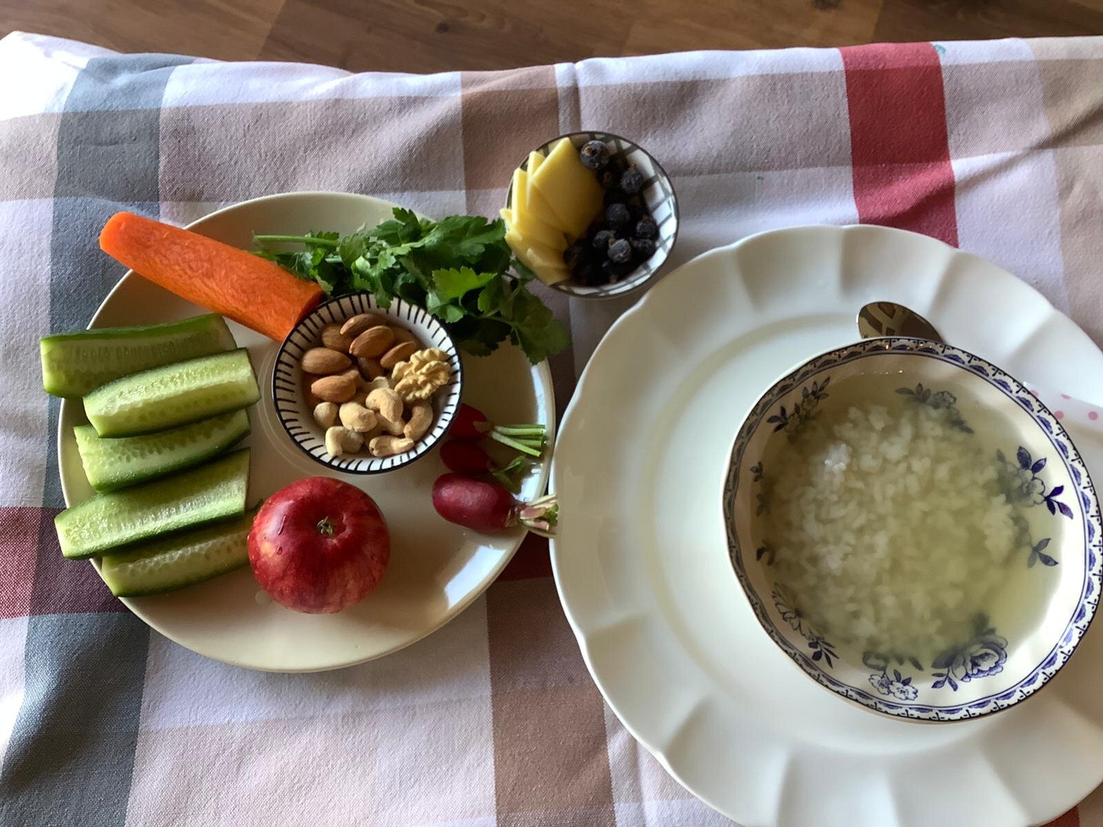 Plate with sliced cucumber, carrot, radishes, apple, mixed nuts; small bowl with pineapple and blueberries; bowl of rice in water on checkered tablecloth.