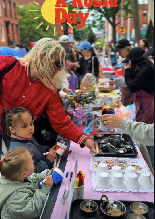 People participating in a community event outdoors, with tables of craft supplies and children in line for activities, under a sign reading 'A Rosie Day' with a yellow sun illustration.