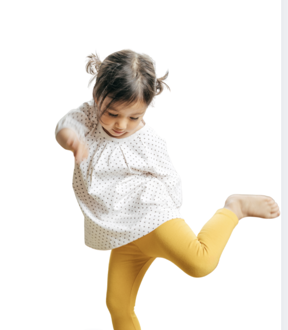 Young girl with brown hair in pigtails wearing a white polka dot blouse and yellow pants, jumping or dancing with one leg raised and arms slightly bent.