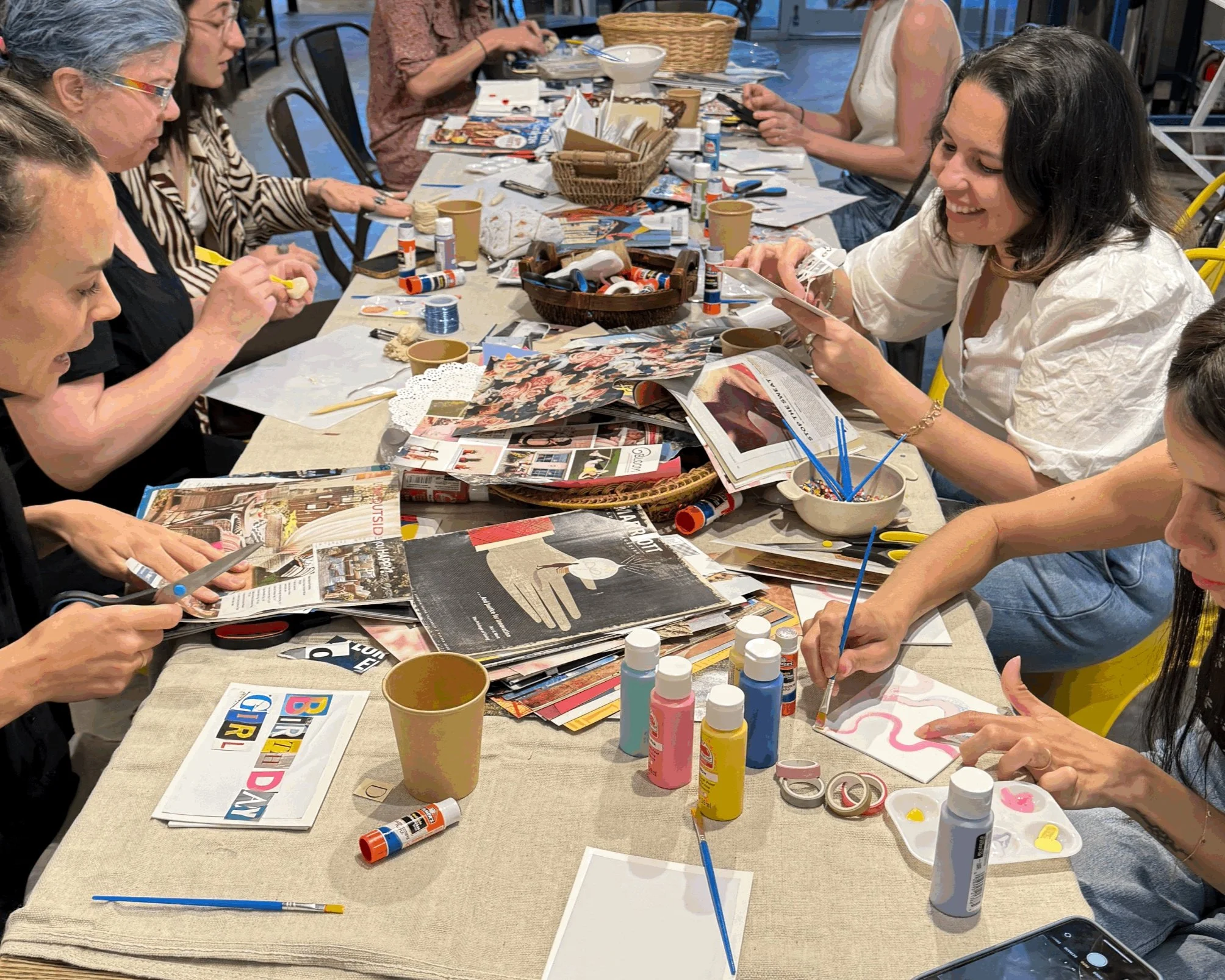 Women decorating cards with paint, glue, scissors, and magazines on a long table.