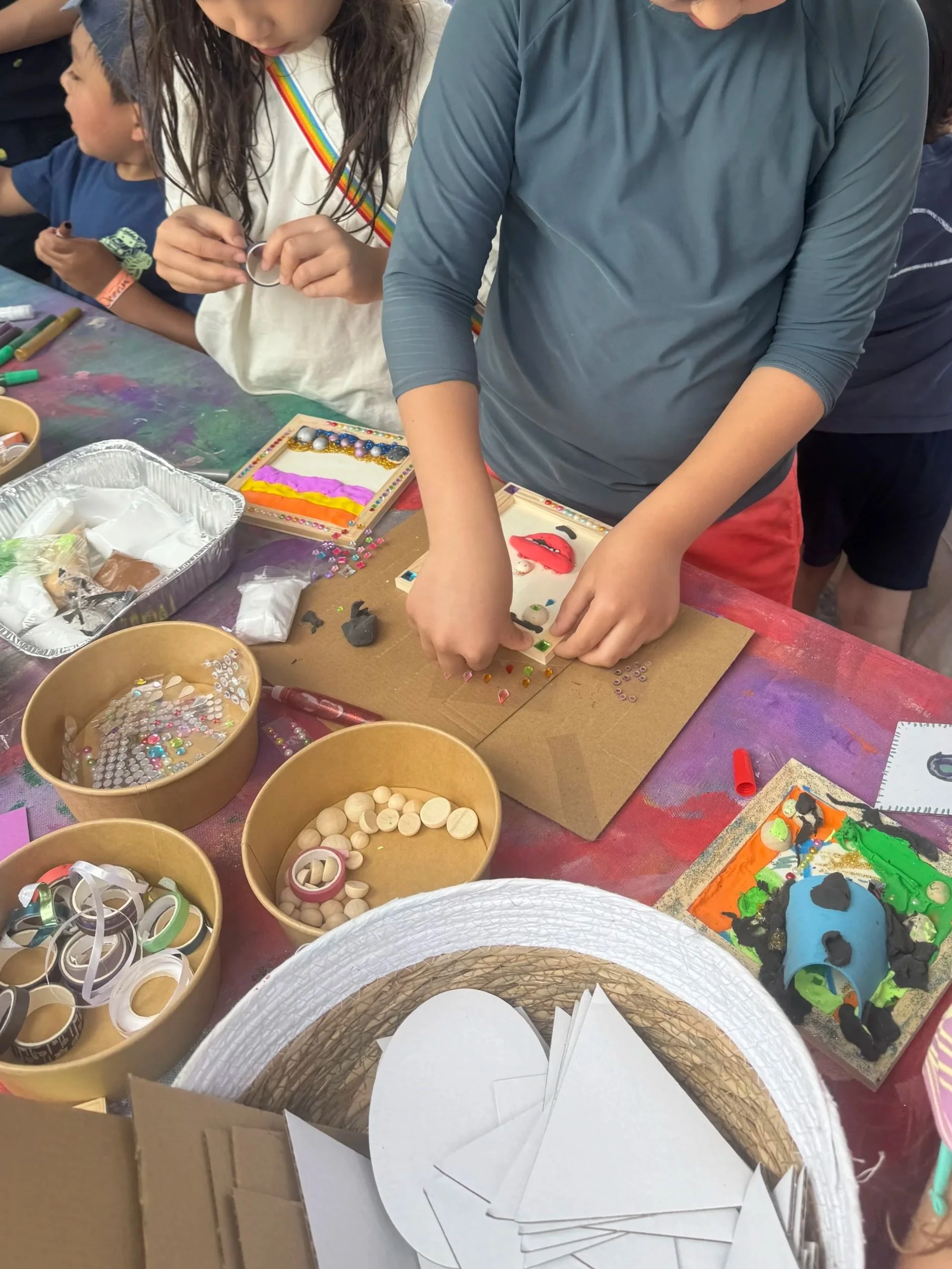 Children and an adult engaged in arts and crafts activities at a table filled with various craft supplies such as paper, beads, buttons, foam shapes, and colorful paints, with some children in the background working on their projects.