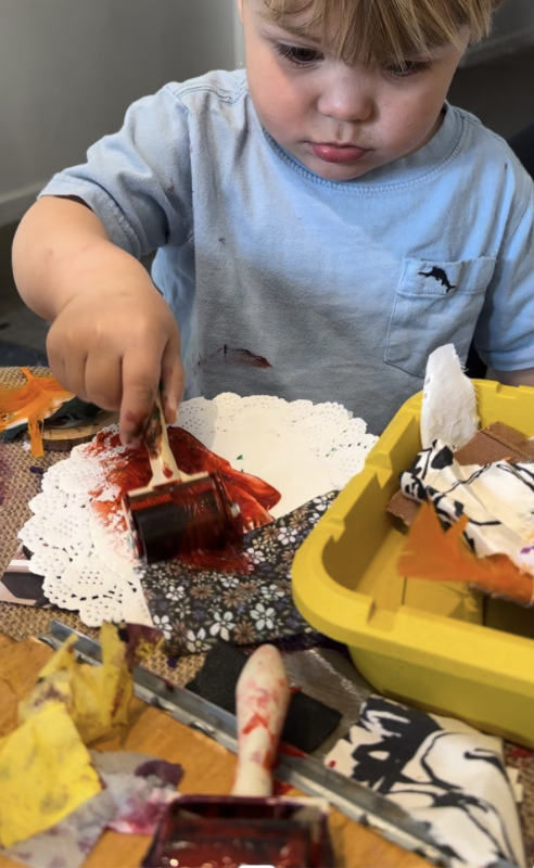 A young boy wearing a light blue shirt is engaging in arts and crafts, using a paint roller to spread red paint on a paper doily on a table with various craft supplies.