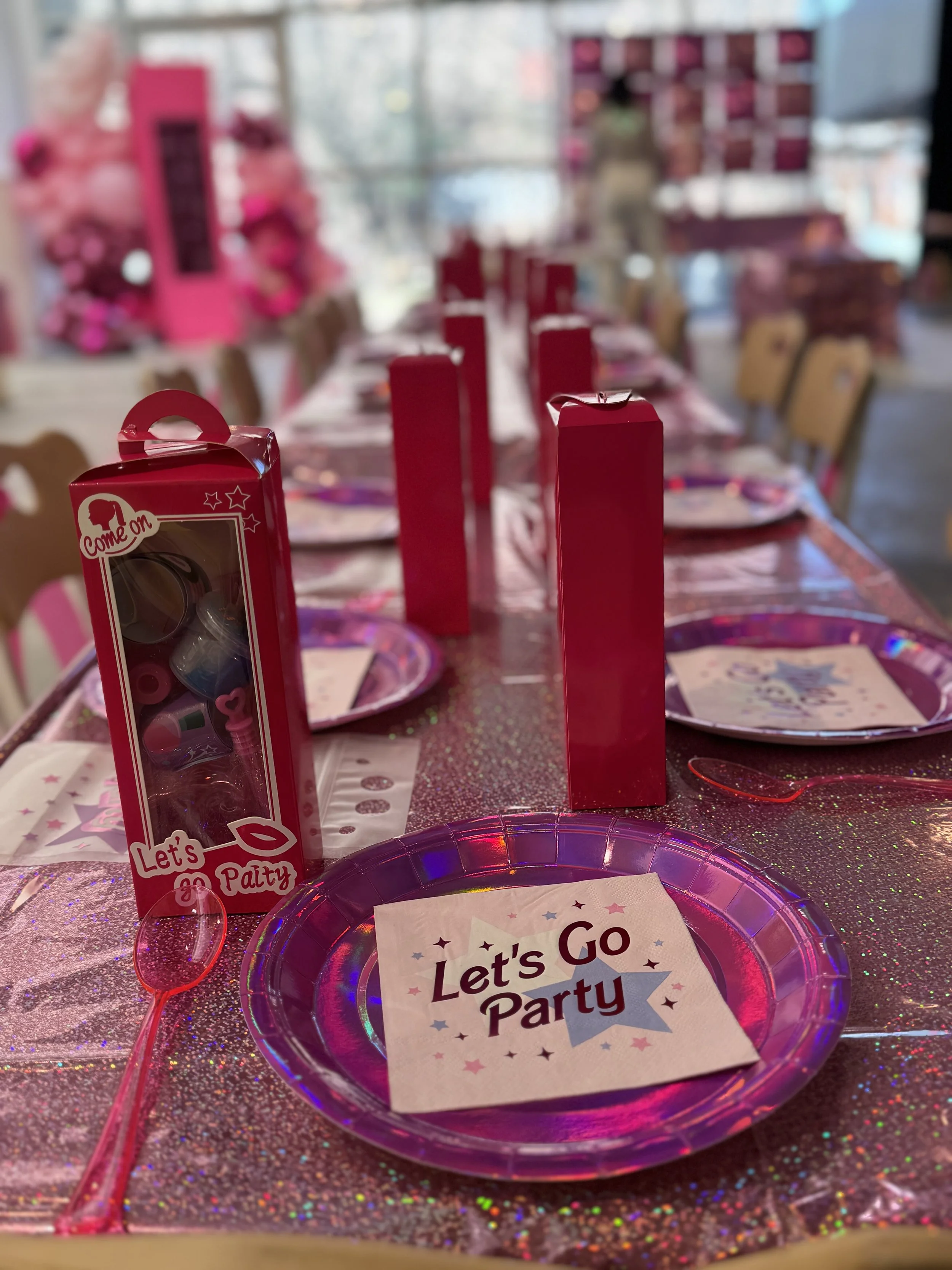 A birthday party table decorated with pink glitter tablecloth, pink party favors, paper plates, paper cups, and a cup cake with a sign that reads 'Let's Go Party' on a pink, holographic plate.