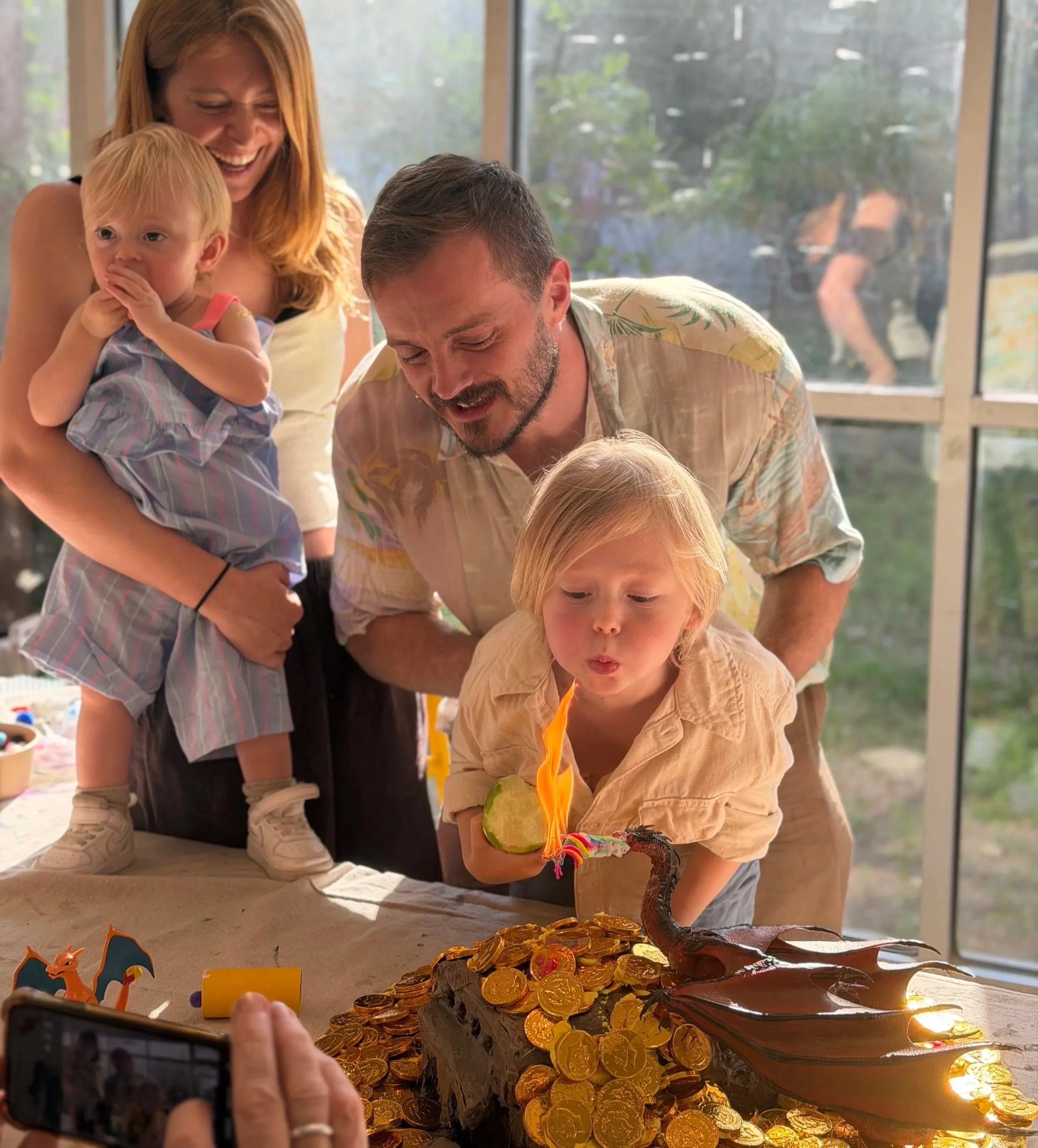 A group of people celebrate a birthday or special occasion with a chocolate cake covered in gold coins and a lit sparkler, with children and adults smiling and taking photos indoors near large windows.
