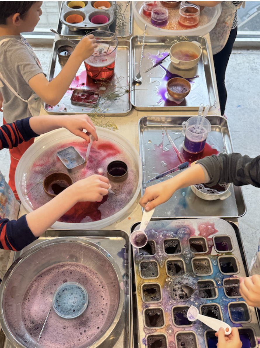 Children making colorful soap or slime in cups and molds at a crafting table with various liquids and tools.