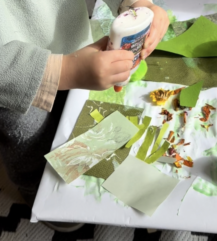Person applying glue to green paper with torn colored paper and floral cutouts on a table for arts and crafts.