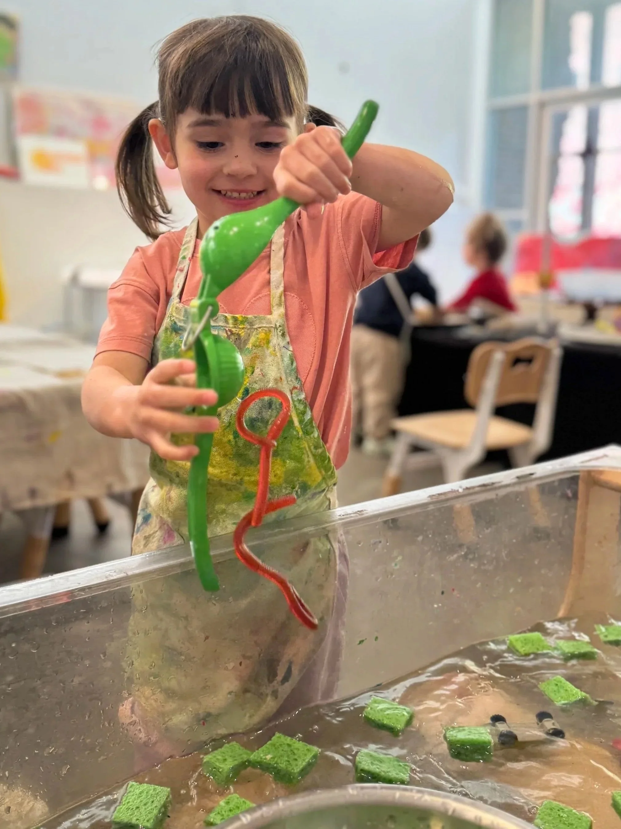 A young girl with dark hair in pigtails, wearing an apron, is flipping green sponge cakes in a pan filled with water or syrup, in a classroom or kitchen setting. Other children are blurred in the background.