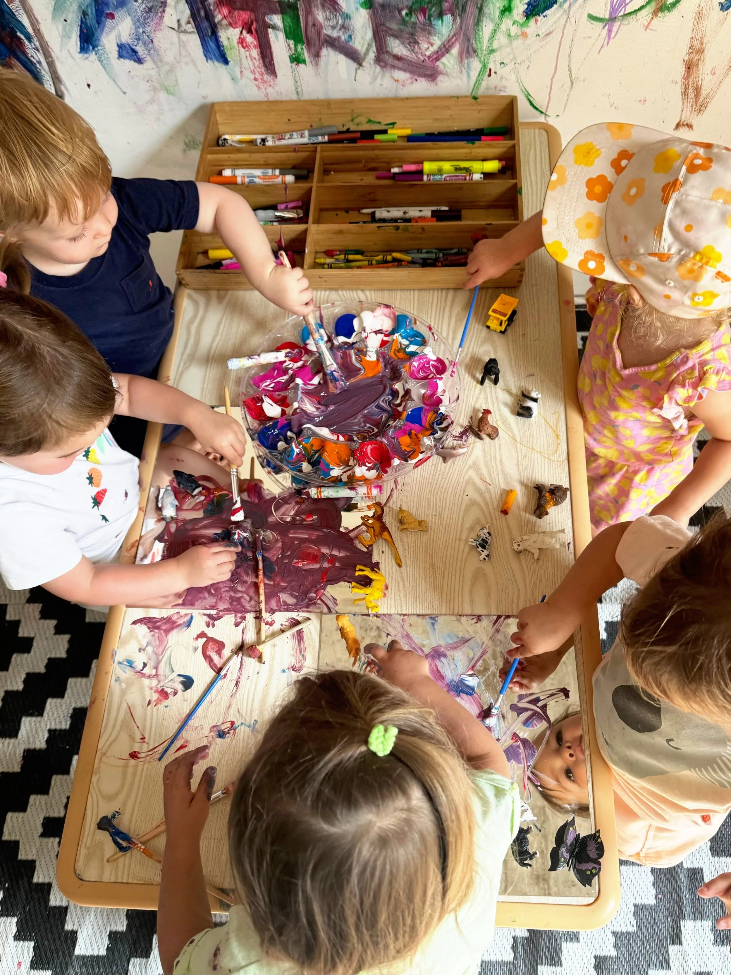 Children engaged in a painting activity at a table, using brushes and colorful paints, with a wooden organizer filled with markers behind them and a wall with colorful artwork in the background.