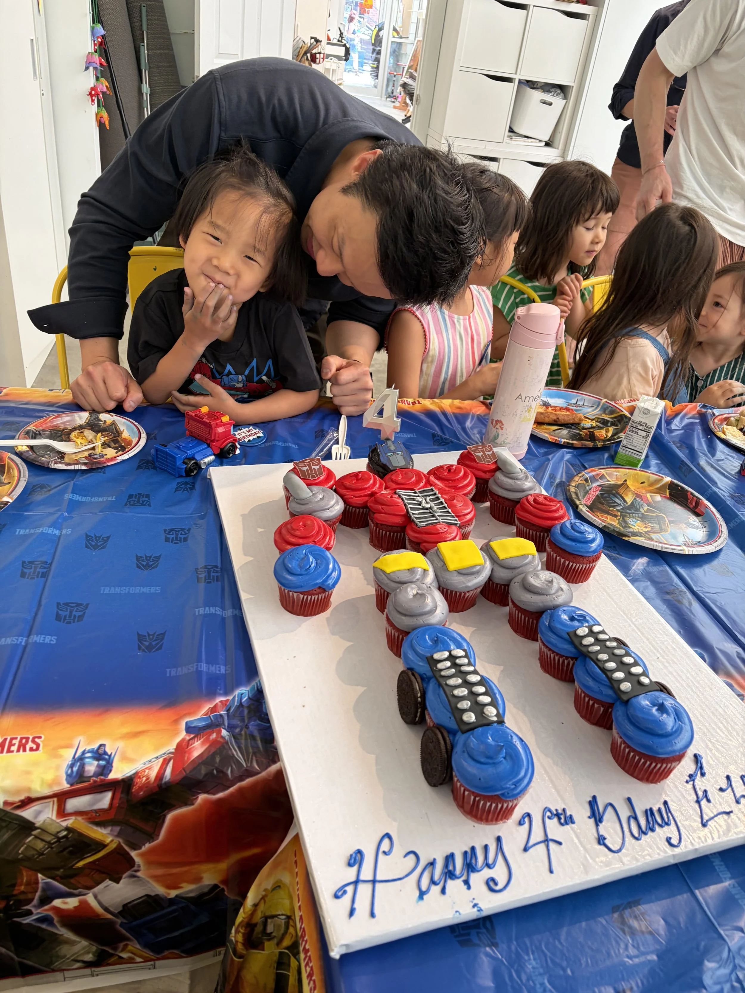 Children and adults gathered around a table with Transformers themed cupcakes and a birthday cake, celebrating a child's birthday.