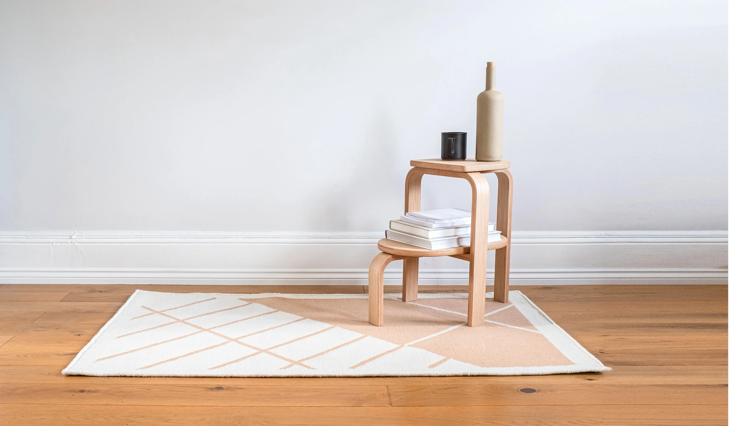 A wooden side table with a black mug, a beige bottle, and stacked white books, positioned against a plain white wall on a hardwood floor with a cream-colored rug.