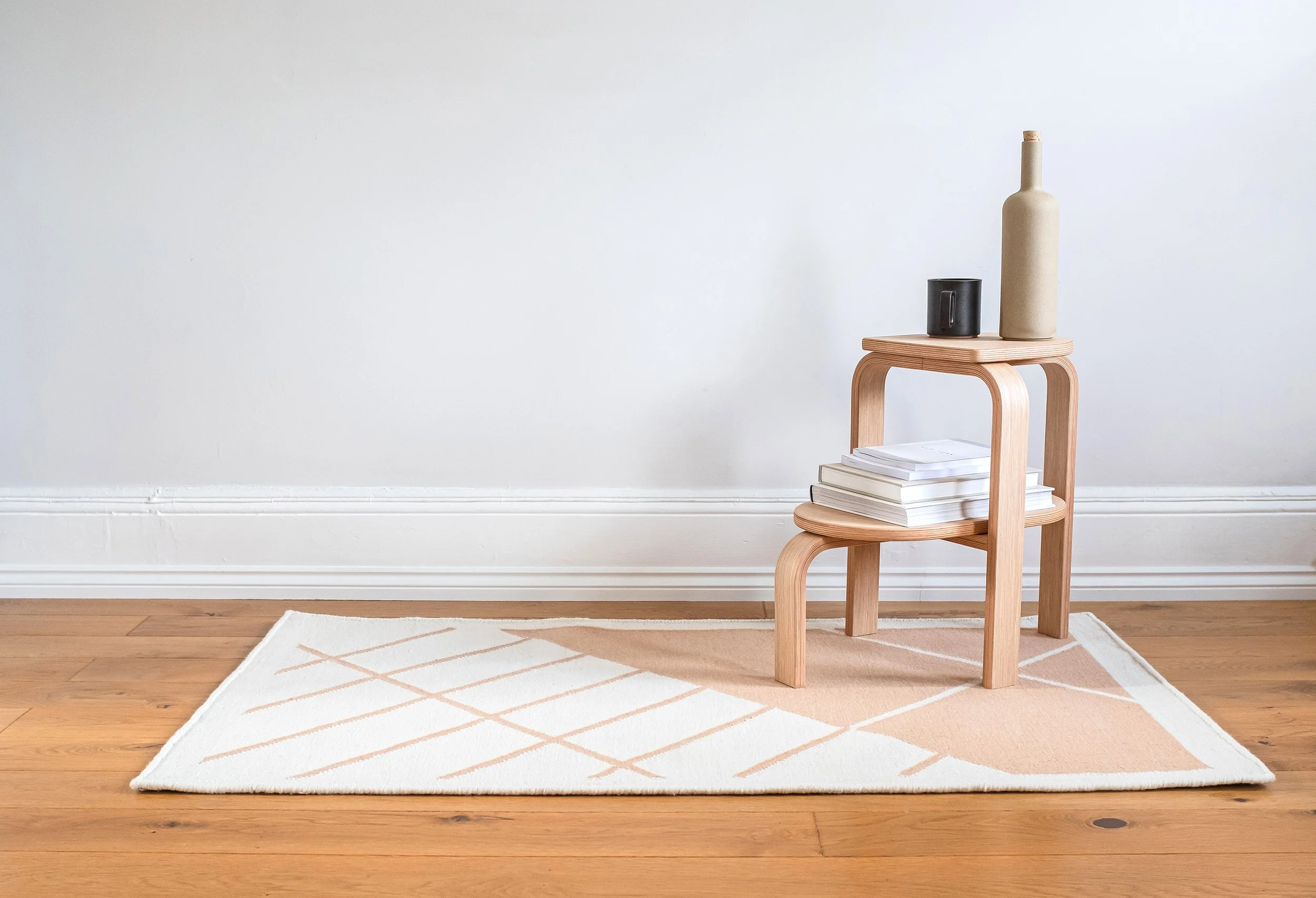 A wooden side table with books at the bottom, topped with a black mug and a beige ceramic bottle, placed on a white area rug with beige geometric patterns on a wooden floor against a plain white wall.