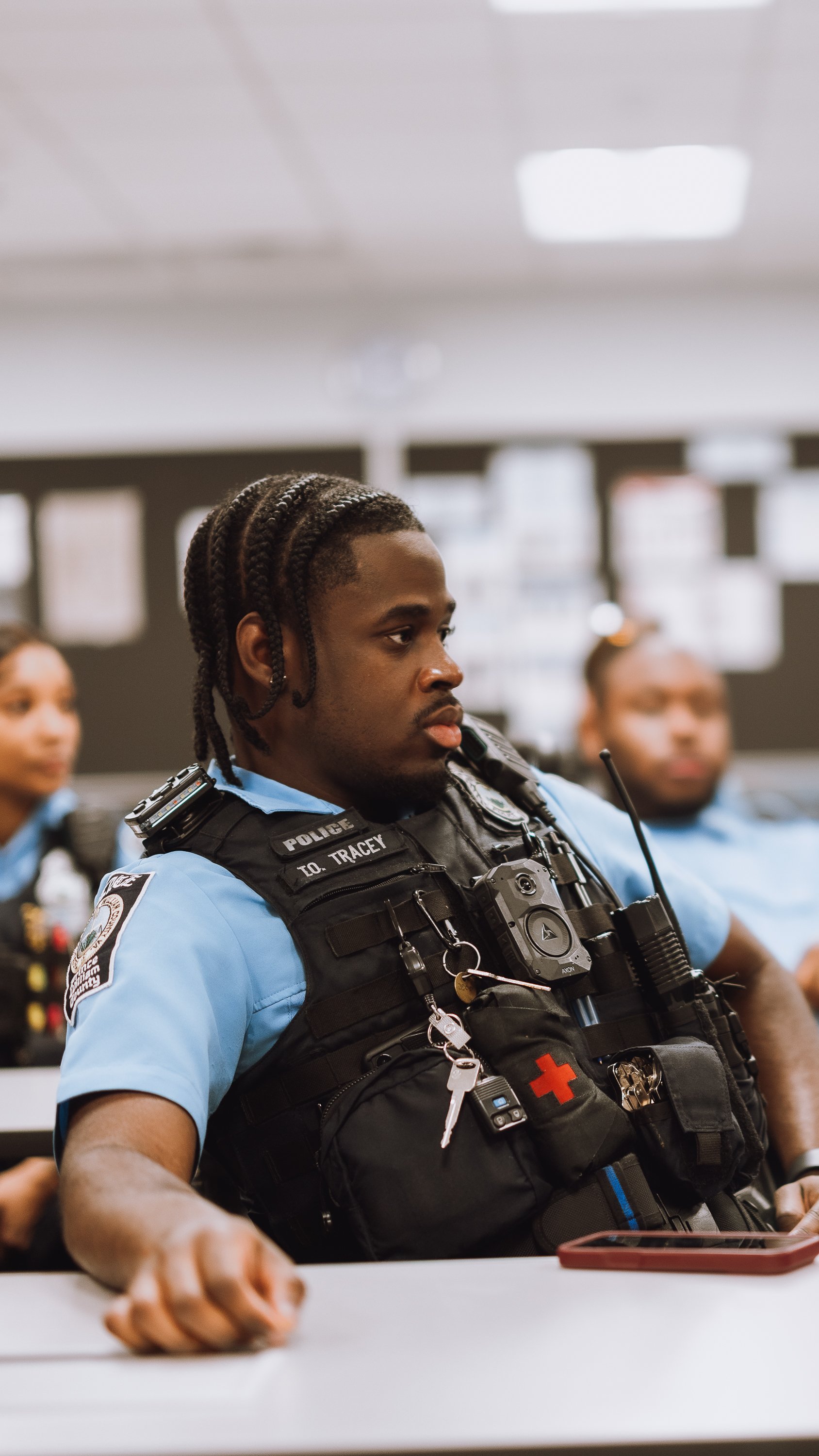 A police officer in uniform sitting at a desk in a classroom or training room, surrounded by other officers, listening attentively.