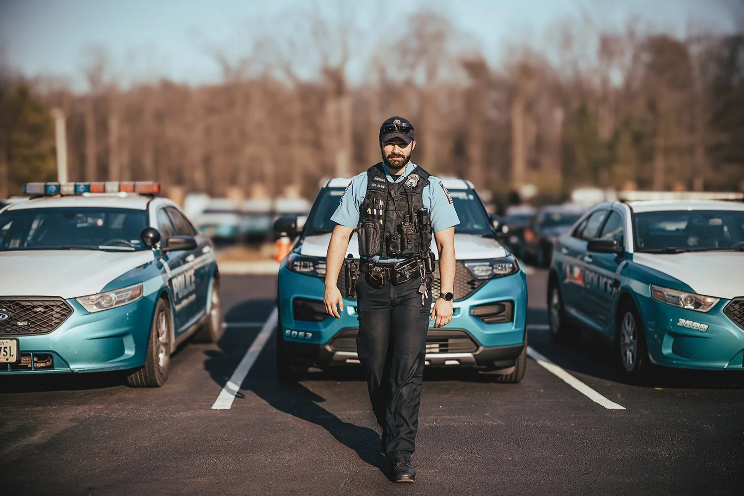 A police officer walking in a parking lot with police cars on either side, some background trees, and a partly cloudy sky.