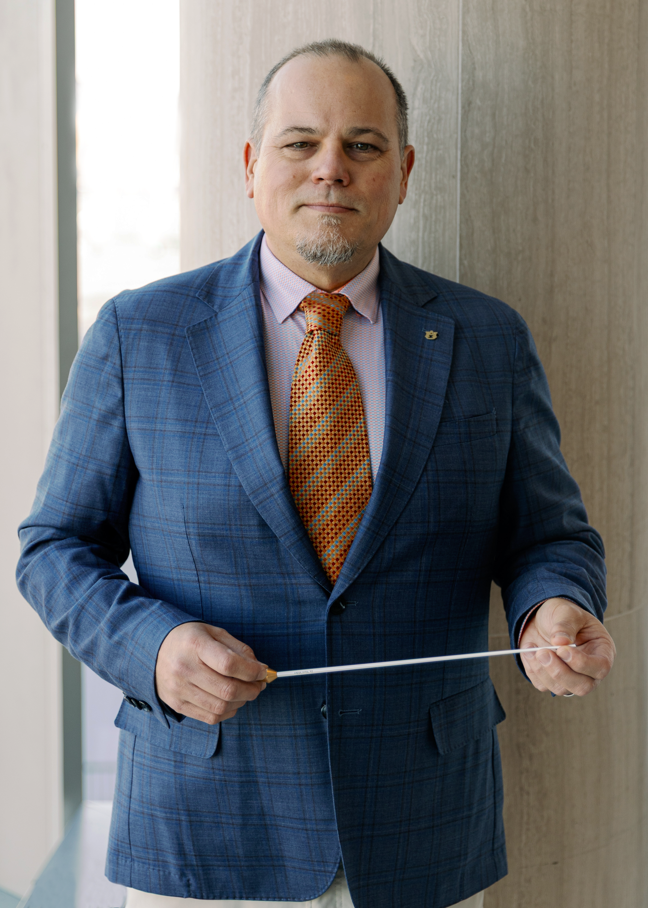 Dr. Ward Miller stands in a blue suit, orange collared shirt, and orange patterned tie standing indoors, holding a white conductor's baton.