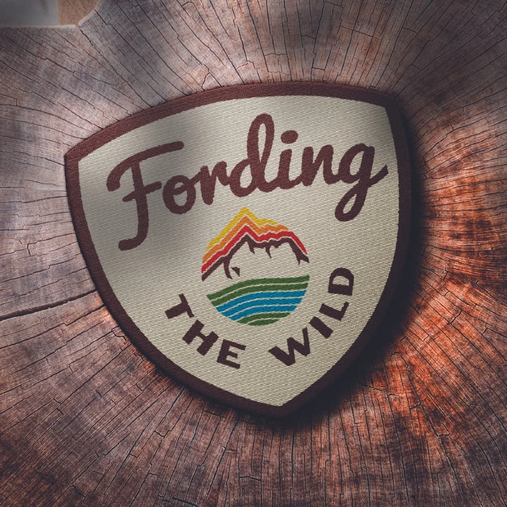 A patch with the words 'Foraging the Wild' and a mountain, river, and sun design on a wooden background.