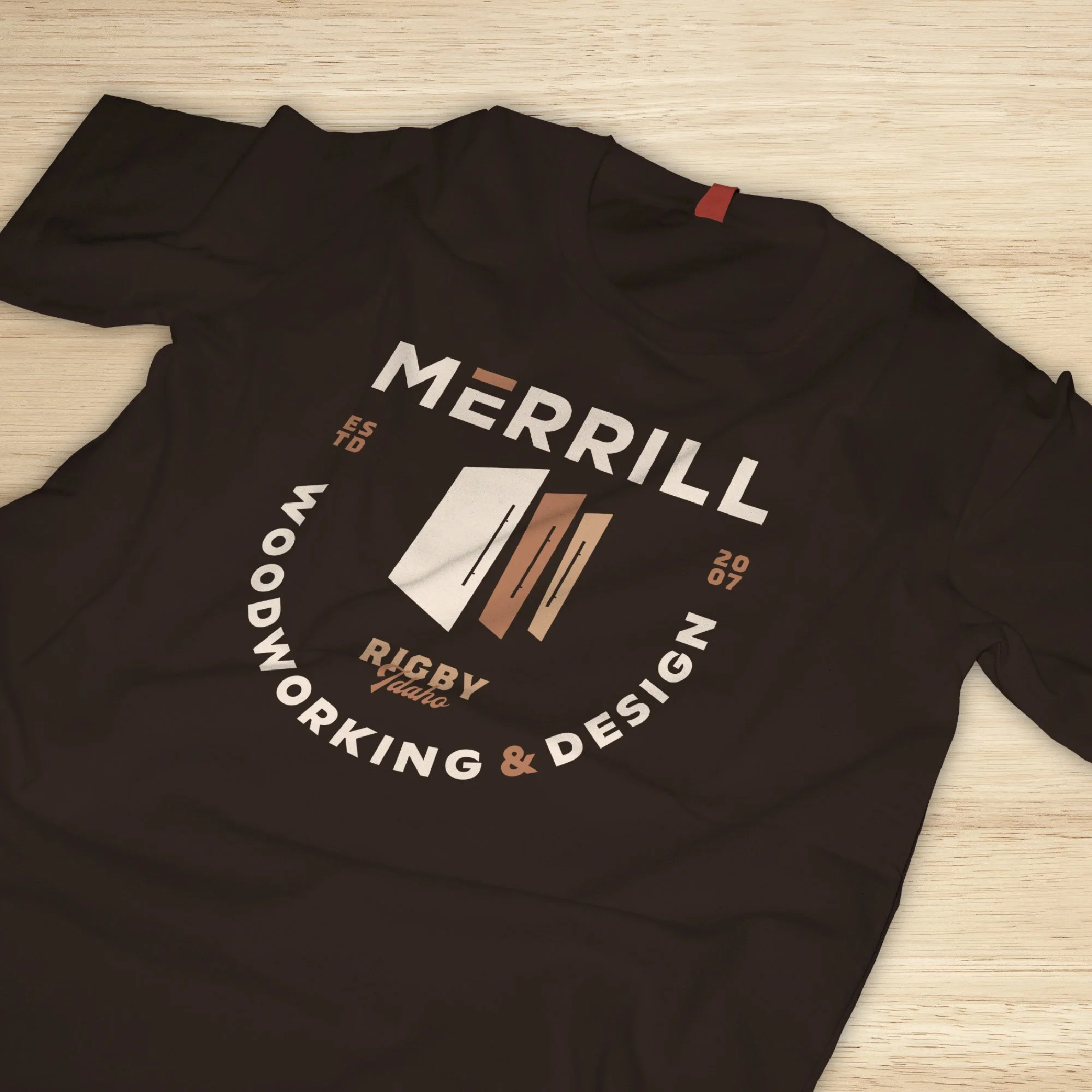 Black T-shirt with circular logo for Merrill Woodworking & Design, featuring a stylized wood pattern and the text 'Merrill Woodworking & Design' and 'Rugby Idaho' in white and brown.
