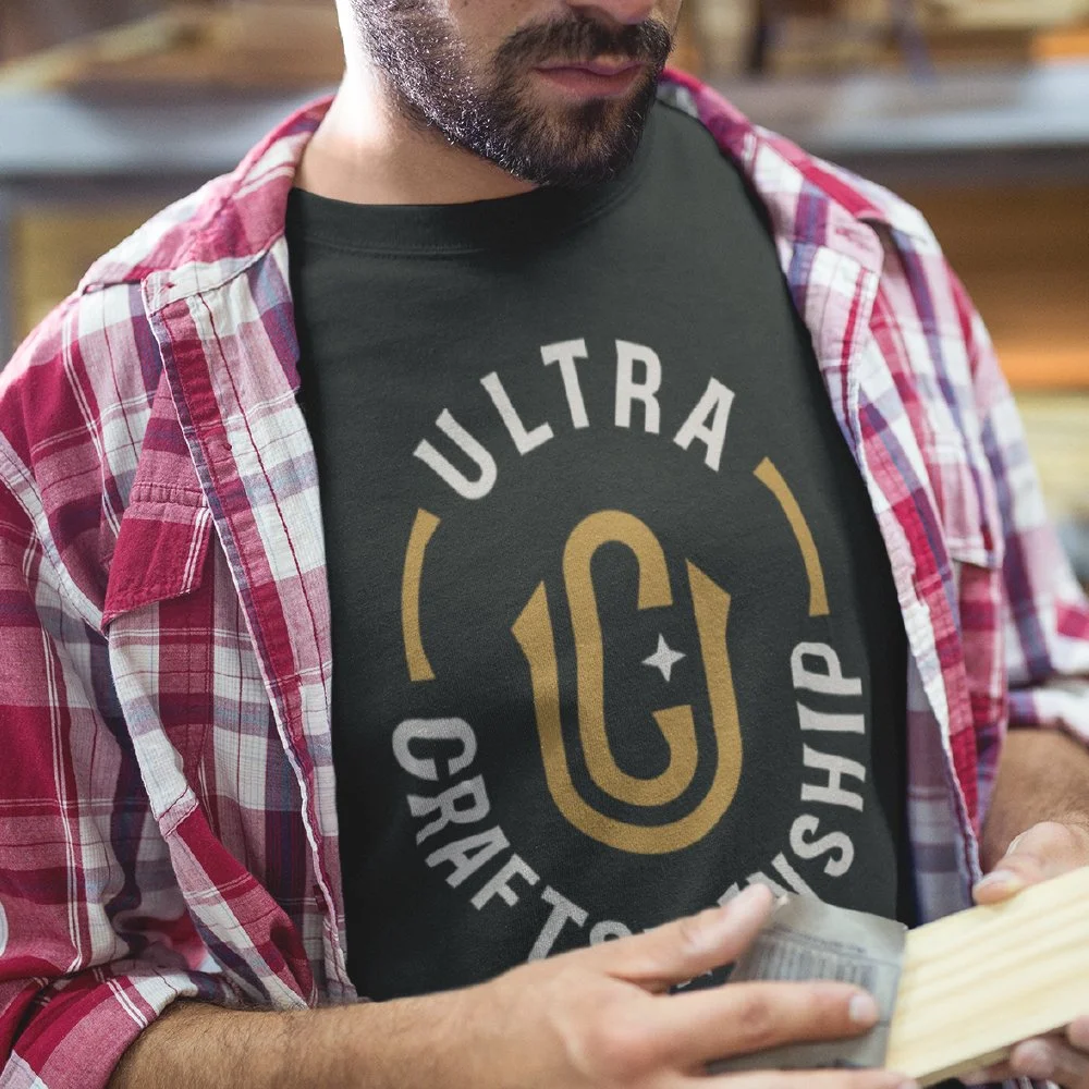 A man wearing a black t-shirt with a gold and white "Ultracraft Camp" logo and a red plaid shirt over it, reading a small book.