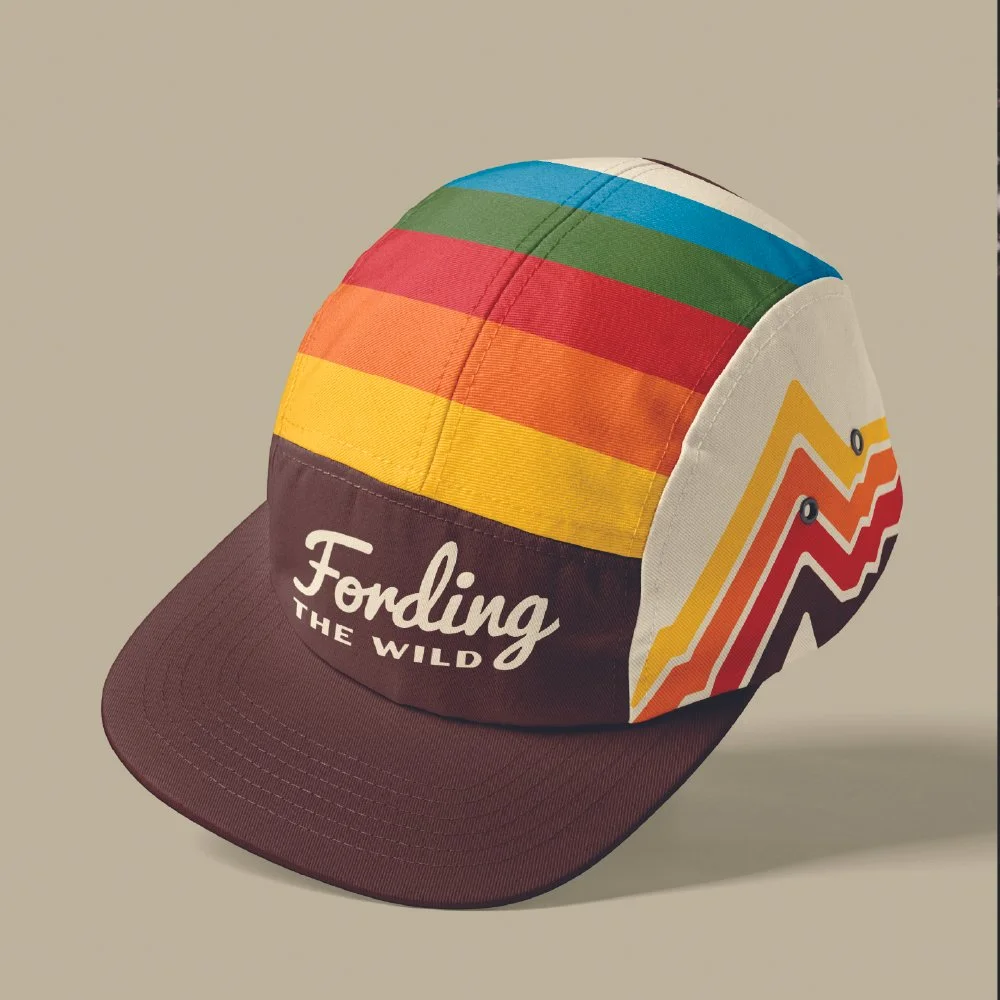 A colorful baseball cap with rainbow stripes on the crown and a mountain graphic on the side, with the text "Fording the Wild" on the front panel.
