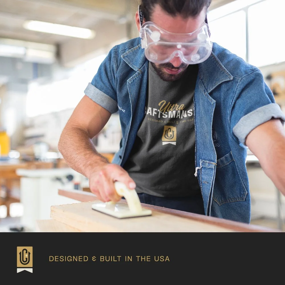 A man in safety goggles and a denim shirt uses a glue gun on a wooden surface in a workshop.