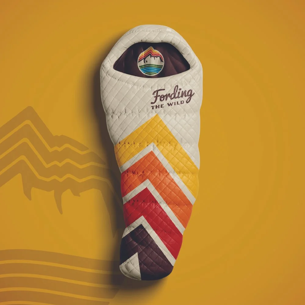 A quilted oven mitt with multicolored chevron pattern, featuring shades of yellow, orange, red, and purple, with the text "Fording the Wild" on it. The mitt is placed on a yellow background with a subtle mountain outline.