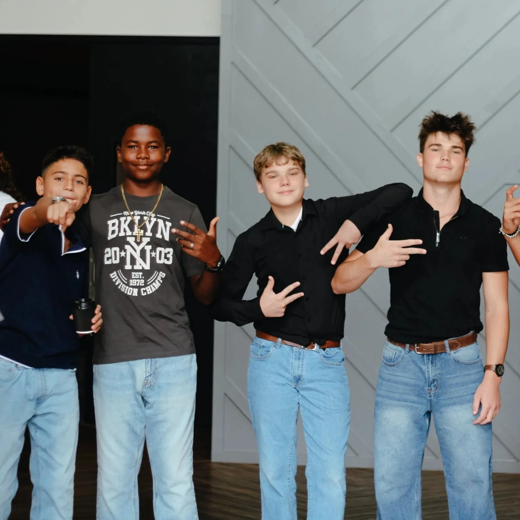 Four young men posing together indoors, wearing casual black or dark-colored shirts and light blue jeans, with one boy hanging a hand with peace signs and the others striking fun poses.