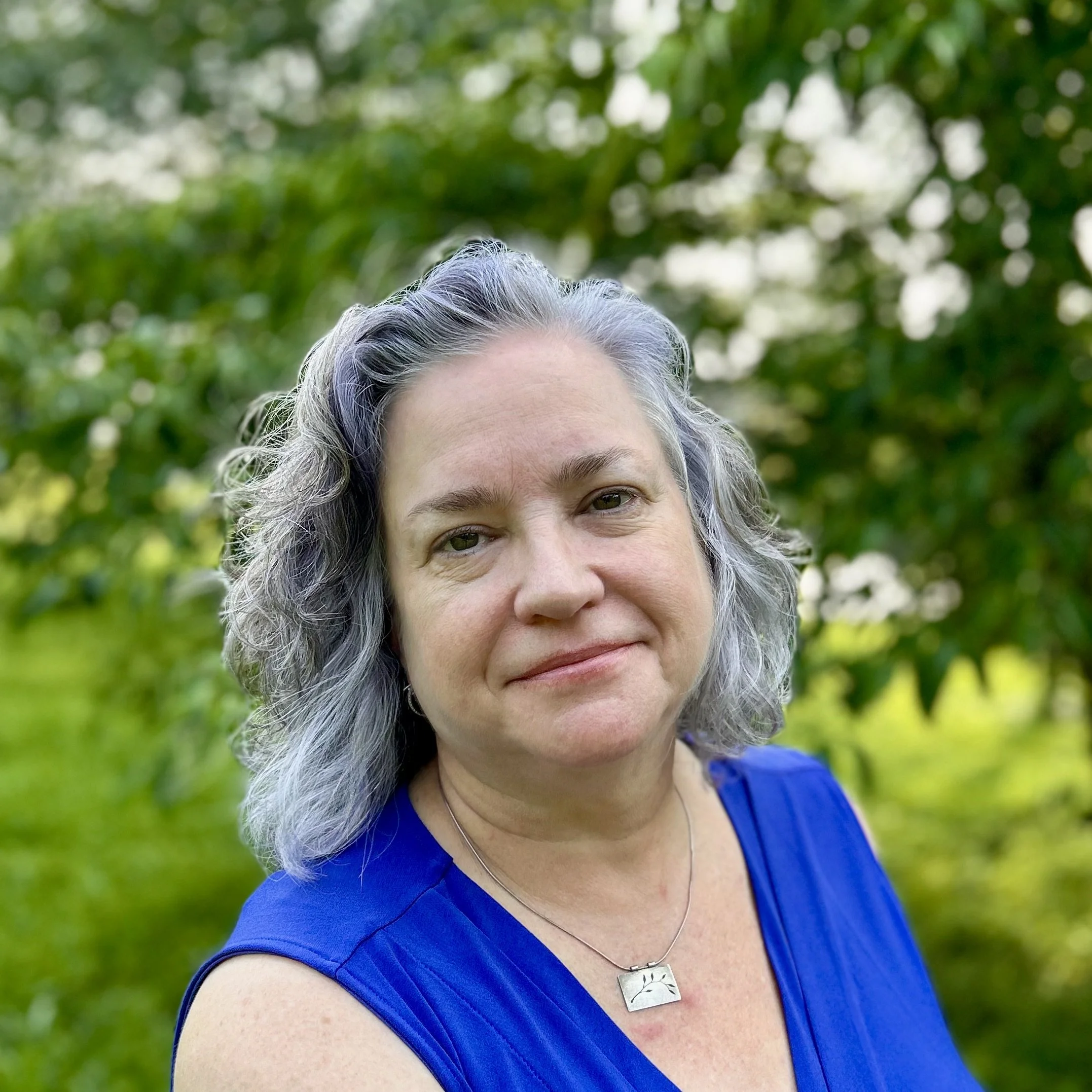 A woman with gray, wavy hair, wearing a black top, silver hoop earrings, and a silver necklace, smiling outdoors with a blurred green forest background.