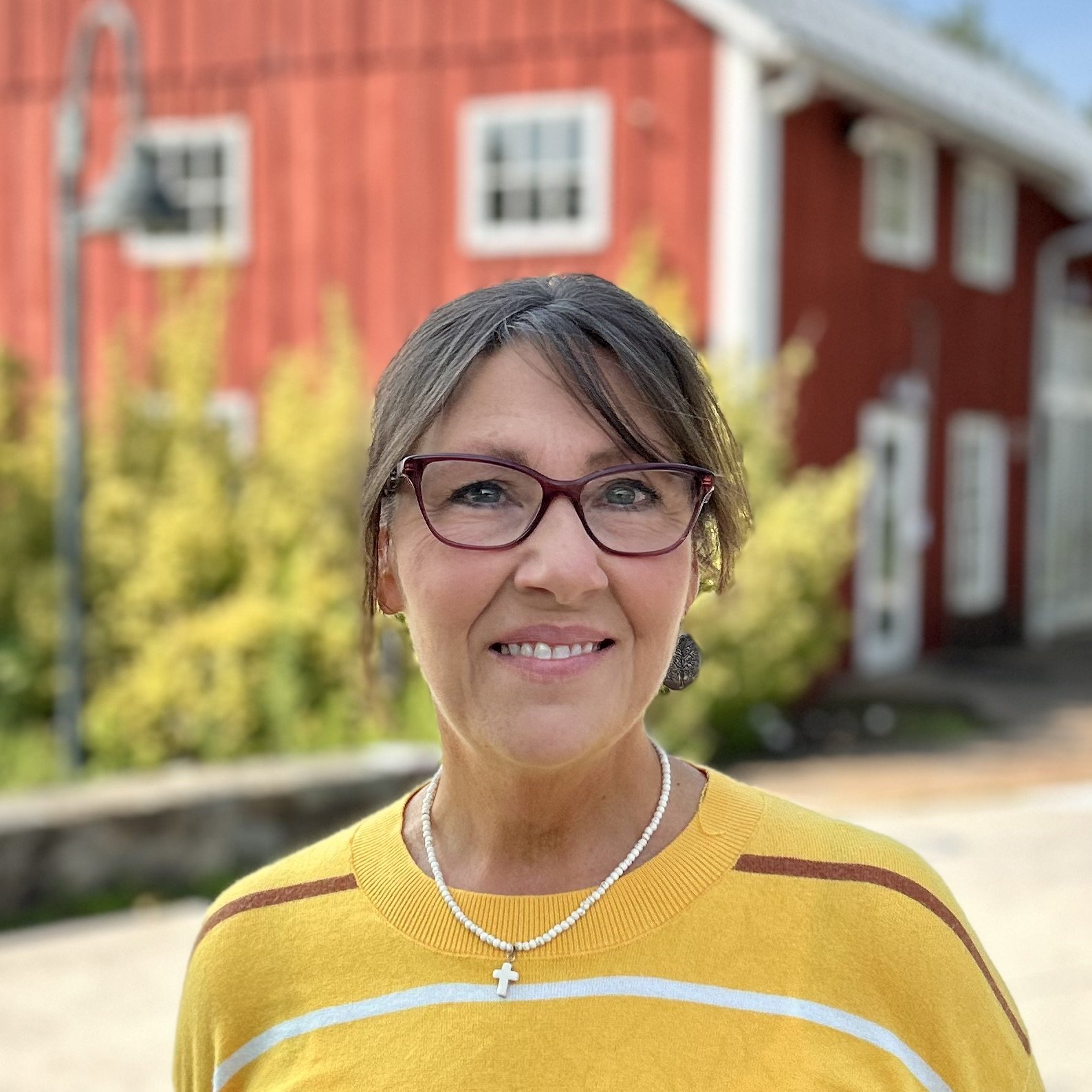 Smiling woman with short brown hair and glasses, wearing a red and white floral top, a pearl necklace with a cross pendant, and large silver earrings, standing outdoors in front of a red barn and green foliage.