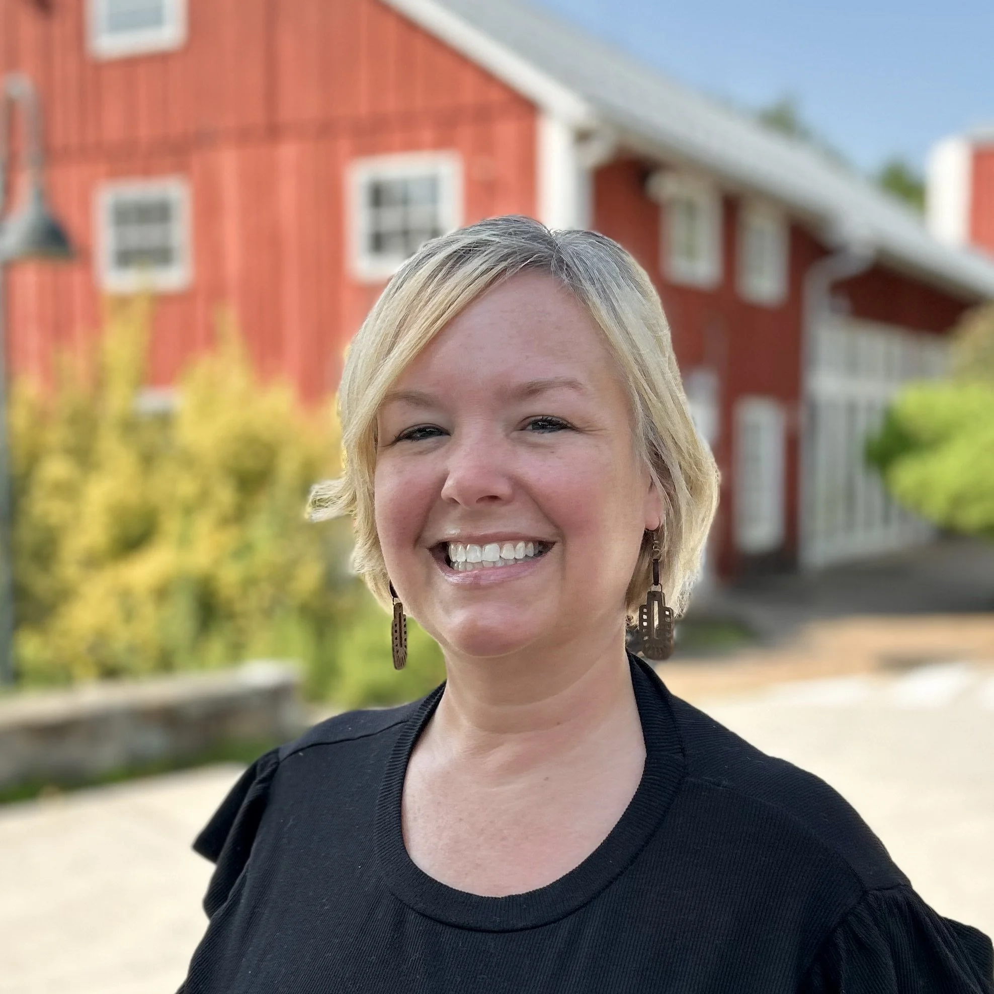A smiling woman with short blonde hair standing outdoors in a park during autumn, wearing an orange cardigan over a black top, with colorful fall foliage in the background.