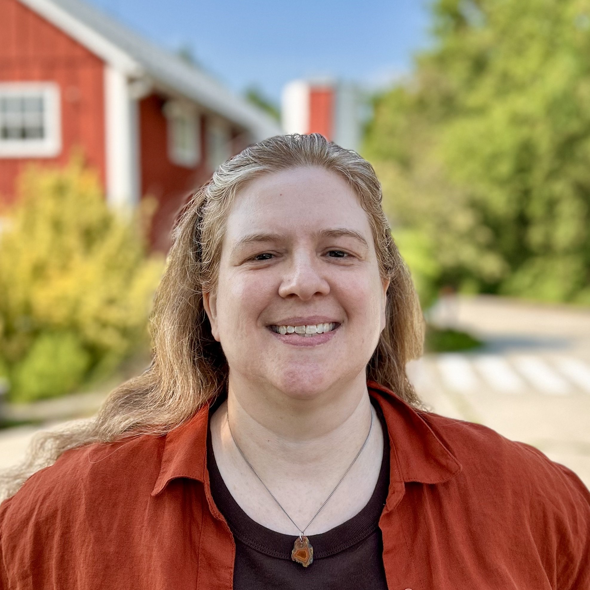 A woman with short blonde hair smiling outdoors, wearing a green jacket and a spiral pendant necklace, with a red building and lush green trees in the background.