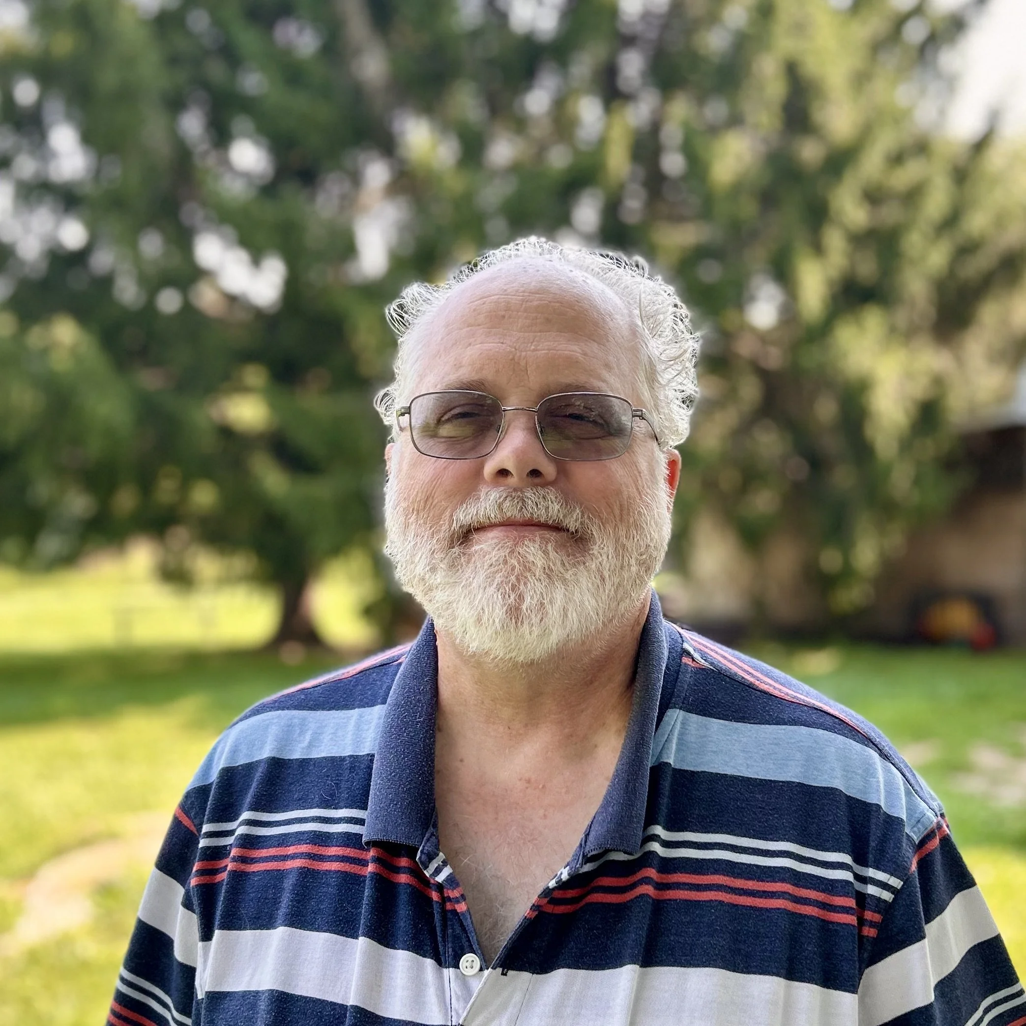 An older man with a white beard, glasses, and a black T-shirt standing outdoors with trees and a cloudy sky in the background.