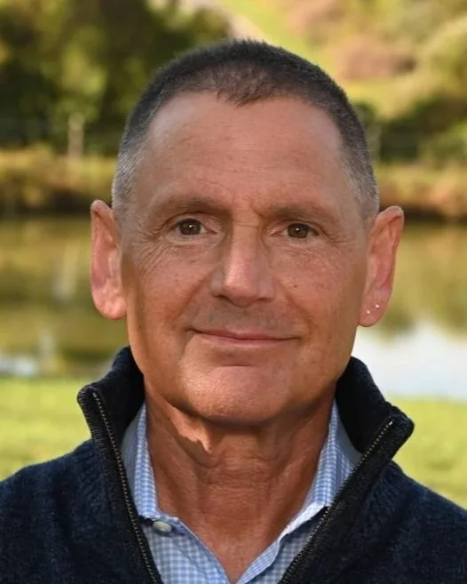 Close-up of a middle-aged man with short hair, wearing a white collared shirt, smiling outdoors near a wall