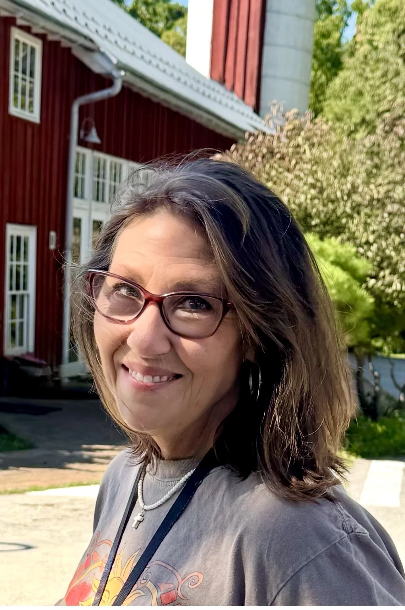 Smiling woman with short brown hair and glasses, wearing a red and white floral top, a pearl necklace with a cross pendant, and large silver earrings, standing outdoors in front of a red barn and green foliage.