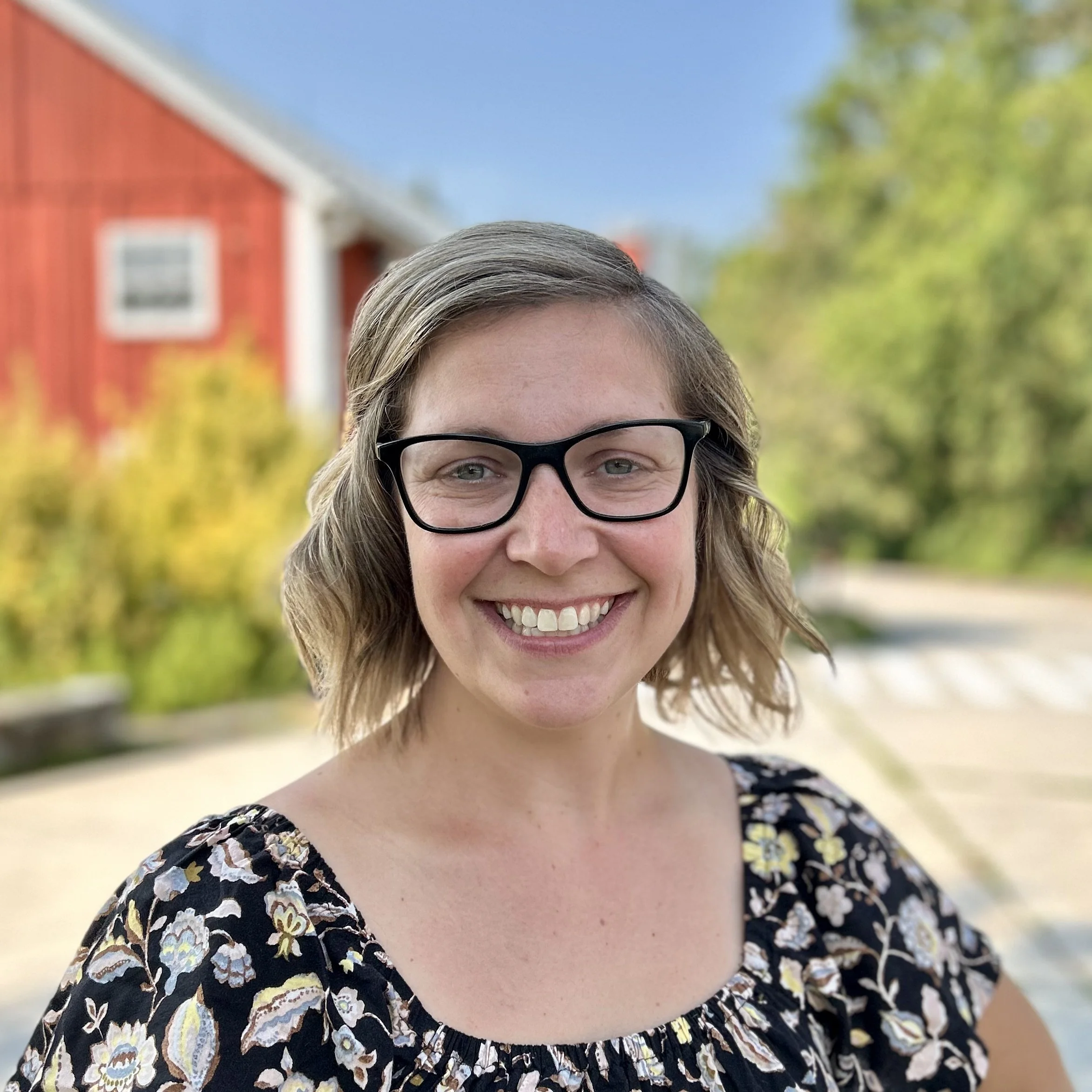 A woman with shoulder-length blonde hair and black glasses smiling outdoors, with a red barn and green trees in the background.