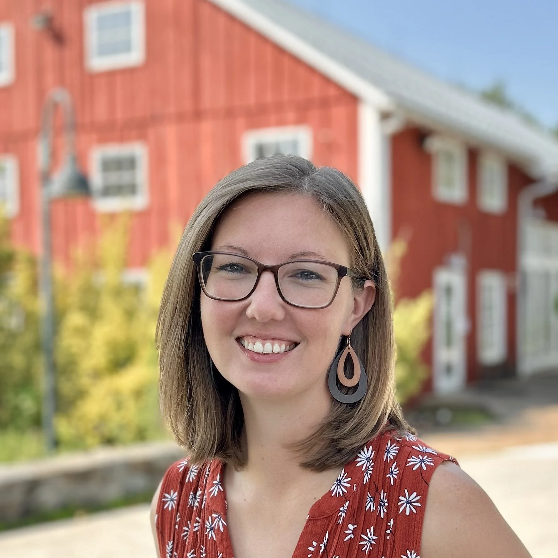 A smiling woman with long wavy hair and glasses standing outside in front of a red barn with green bushes and a clear sky.