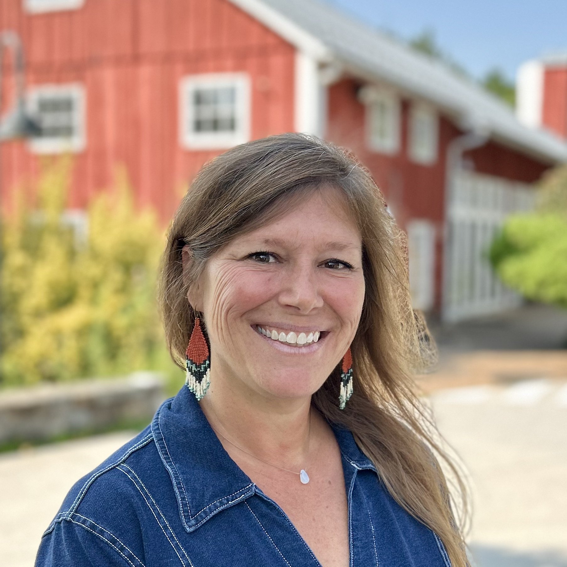 A smiling woman with light brown hair, wearing a maroon plaid shirt and a yellow bandana, standing outdoors near a red building with white-trimmed windows and green trees.