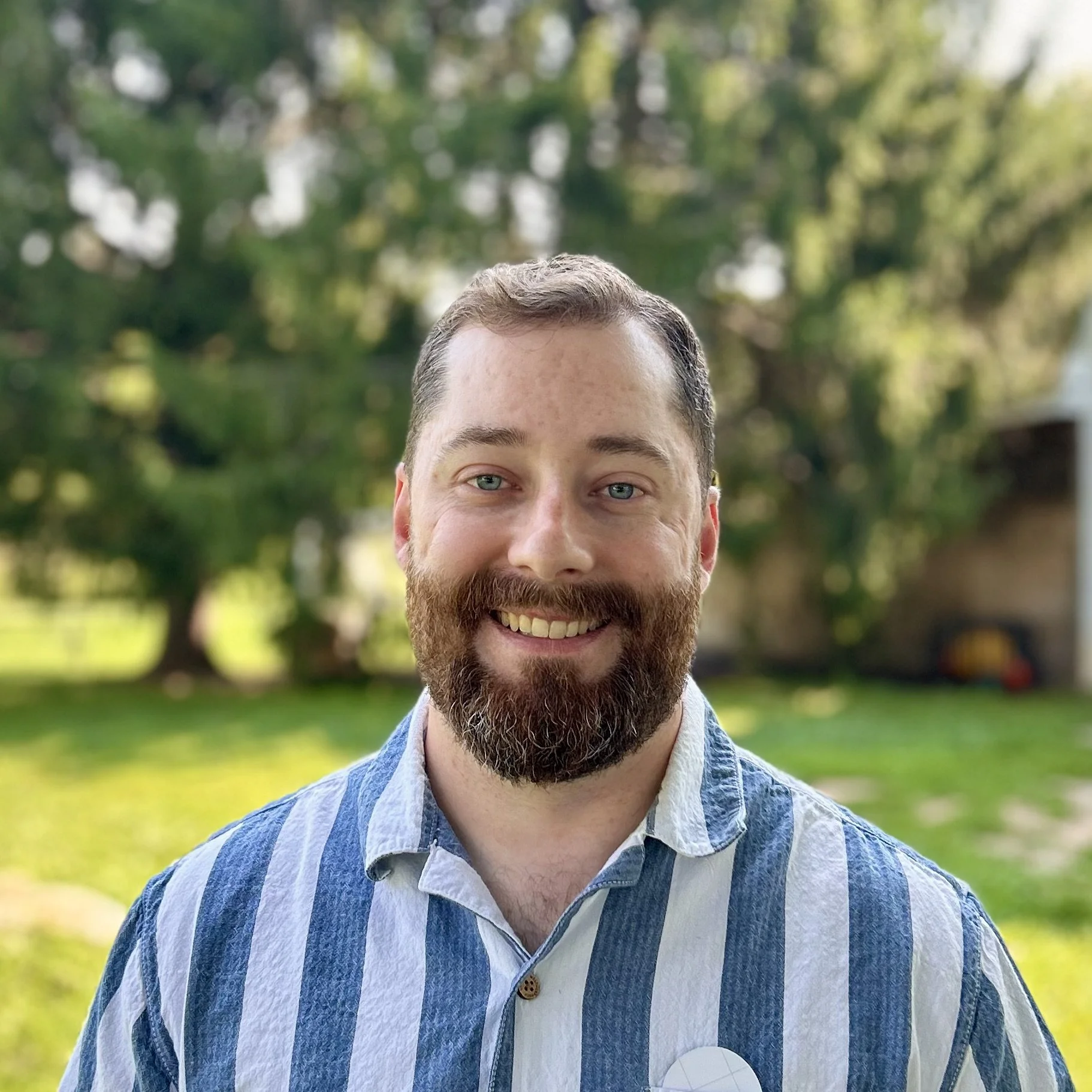A smiling man with a beard and mustache, wearing a dark striped shirt, standing outdoors with green trees and blurred background.