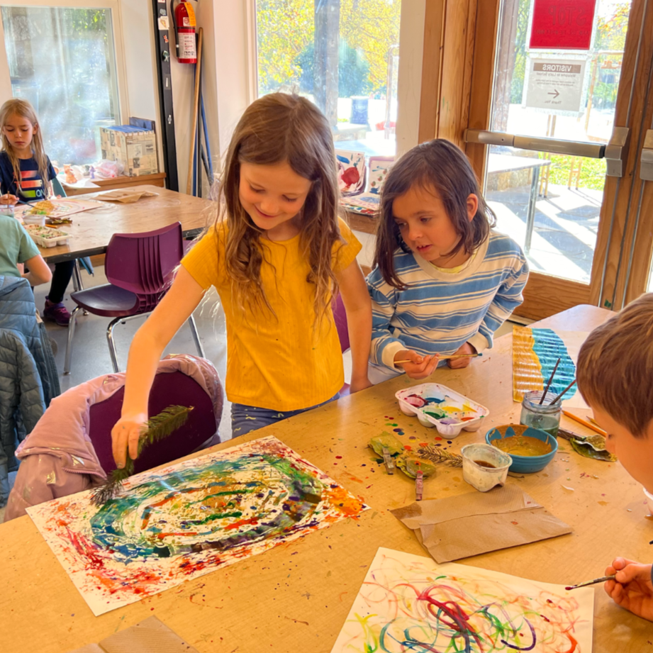 Children in a classroom or art studio engaged in painting and drawing activities, with art supplies on the table.