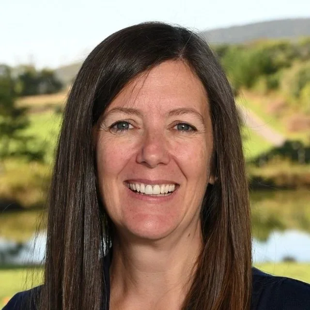 A woman with long brown hair smiling in front of a window overlooking a water body and trees.