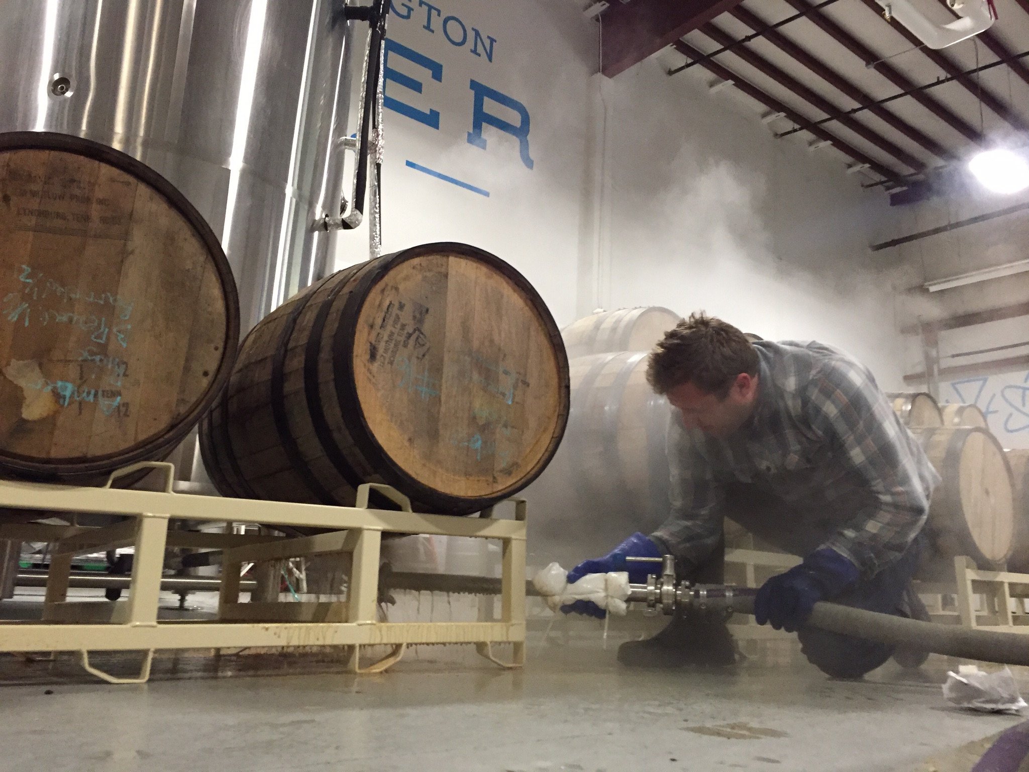 A person works with a hose and fittings beside oak barrels inside the brewery.