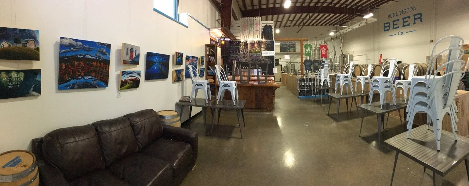 The early taproom with a couch, wall artwork, small bar, tables and brewery equipment beyond.