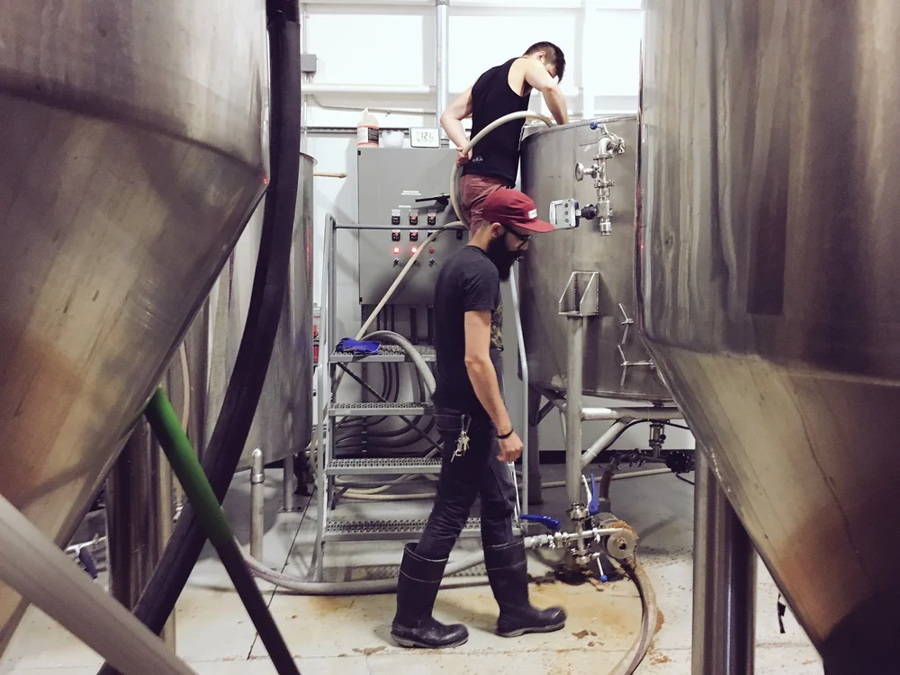 Two people working between tanks on the Bring Forth the Mollusk collaboration brew day at the original Williston brewhouse.