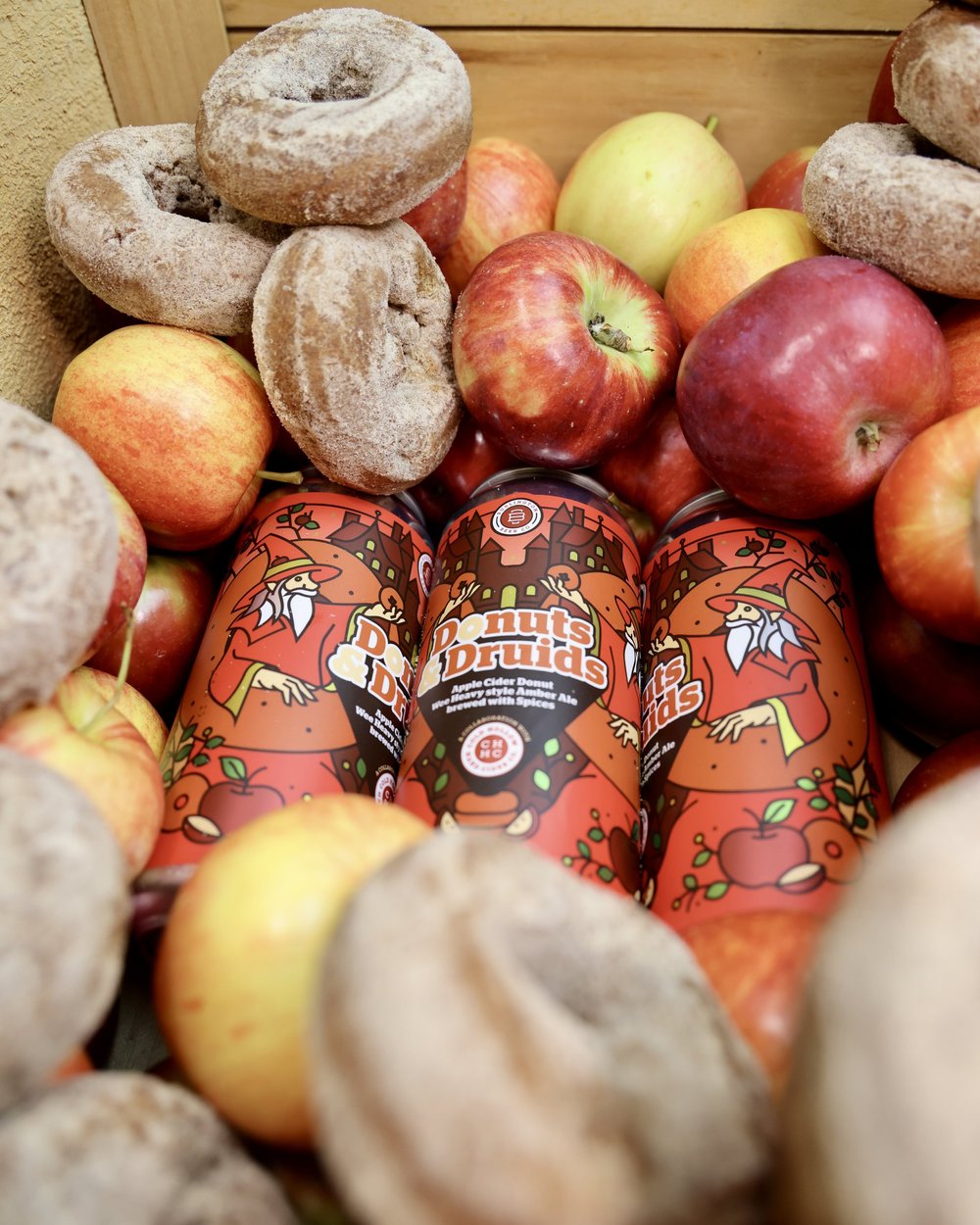 Three illustrated Donuts & Druids cans surrounded by apples and cider donuts.