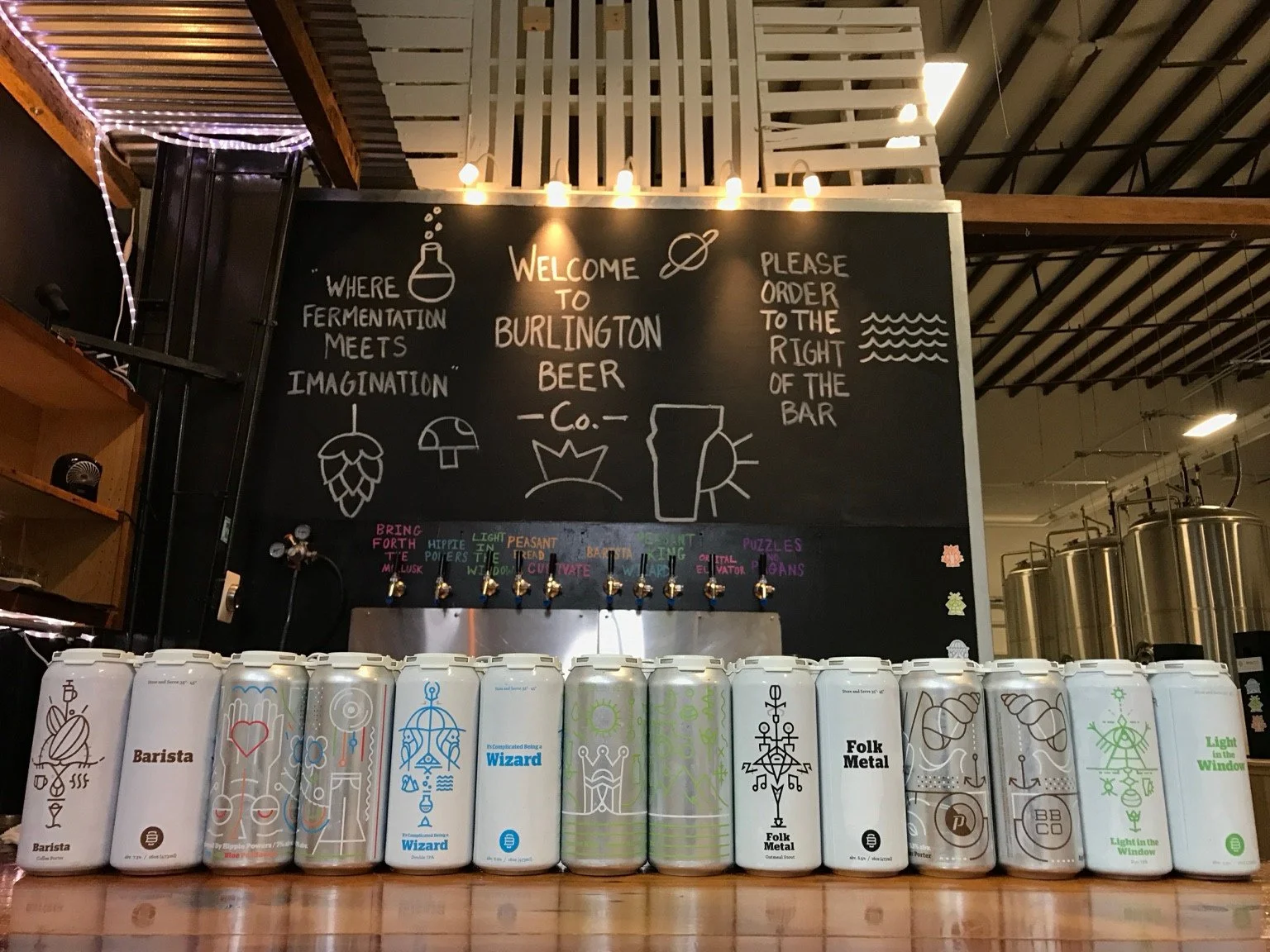 Early Burlington Beer Co. cans lined up below an illustrated chalkboard at the taproom bar.