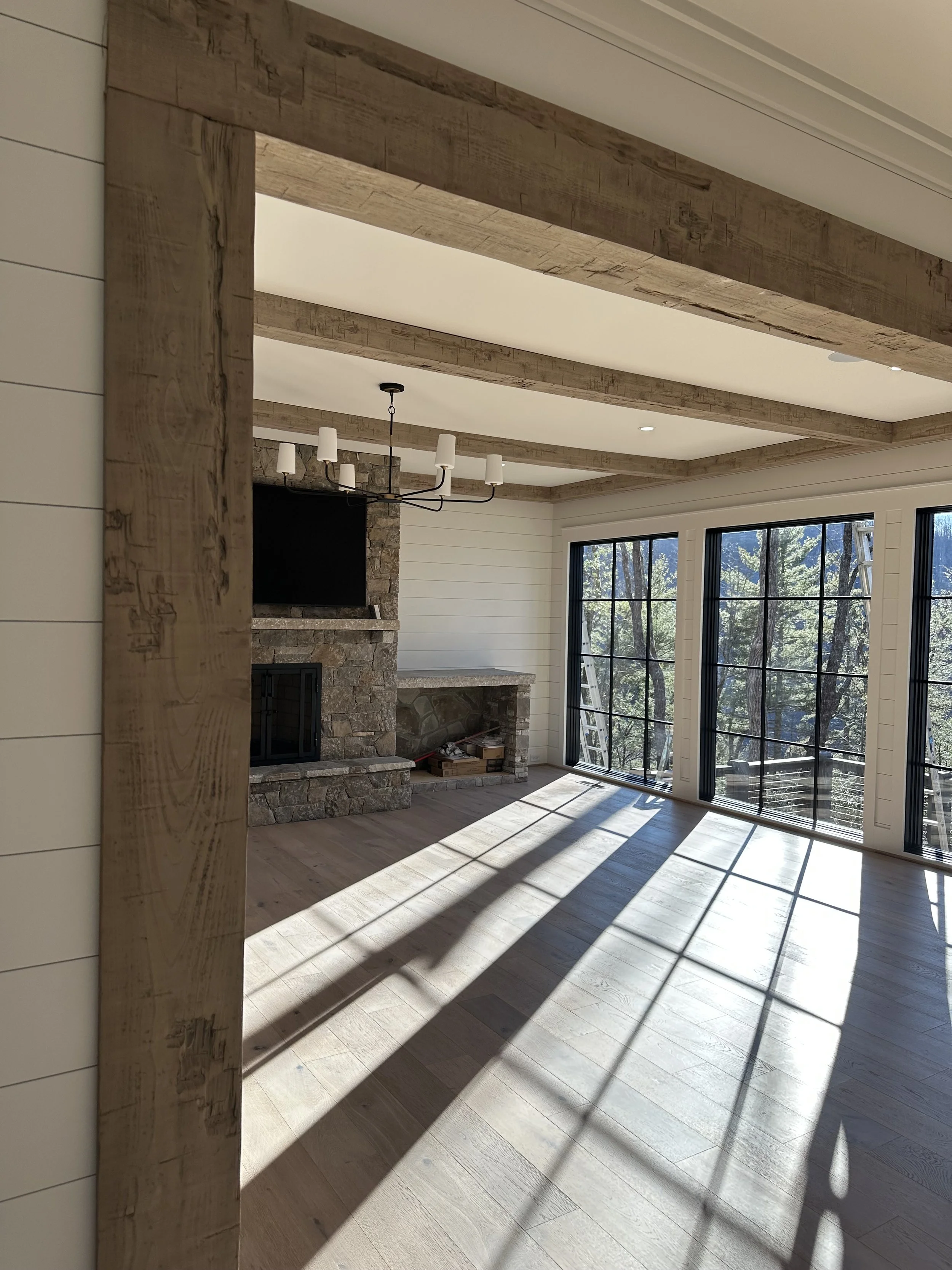 Empty living room with large windows, stone fireplace, modern chandelier, and wooden beams on the ceiling, sunlight streaming through the windows.